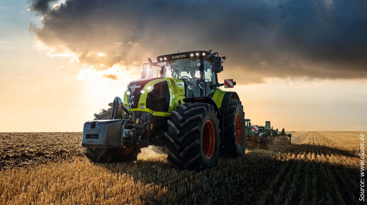 Traktor mit Anhänger der Firma Claas fährt auf einer Straße in einem landwirtschaftlichen Gebiet